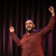A man in a maroon shirt gestures with raised arms and open hands while speaking or performing on a stage with a dark curtain backdrop.