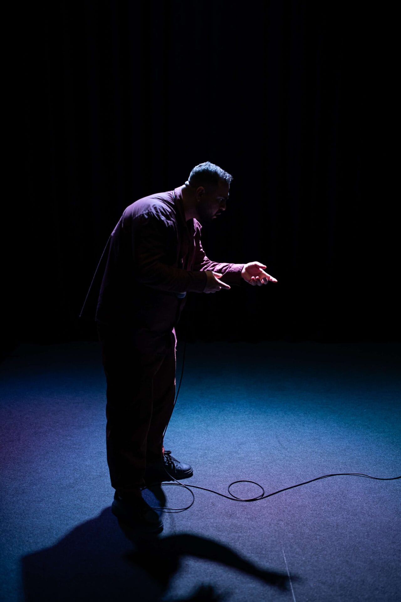 A man stands under a spotlight on a dark stage, holding his hands out and facing downward, with a microphone cable on the floor in front of him.