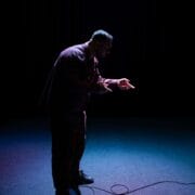 A man stands under a spotlight on a dark stage, holding his hands out and facing downward, with a microphone cable on the floor in front of him.