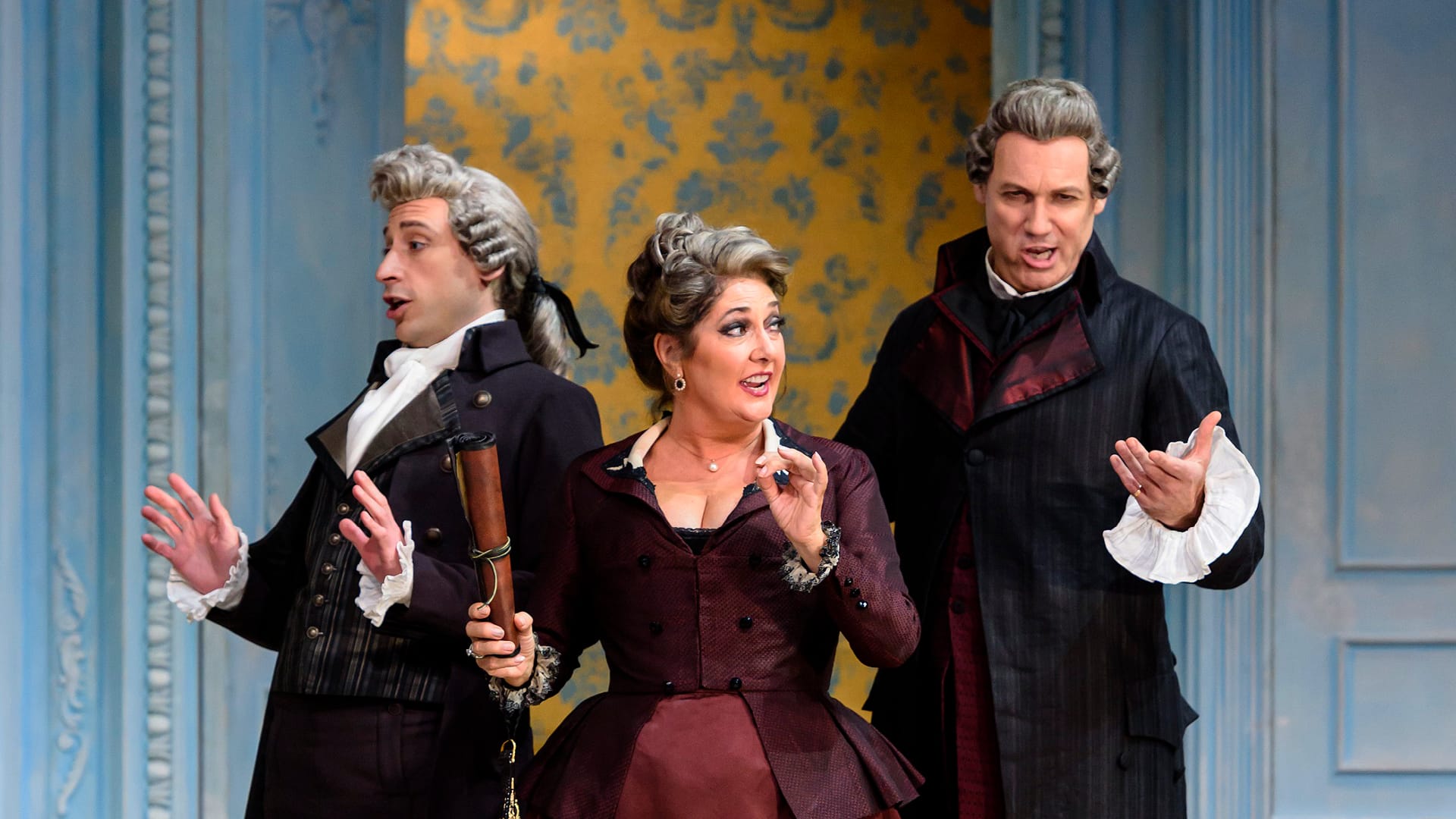 Three people in 18th-century costumes perform on stage, with expressive gestures, in front of a blue and yellow patterned backdrop.