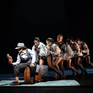 Six dancers in theatrical costumes perform in a line on stage, with suitcases as props, under a focused spotlight against a dark background.