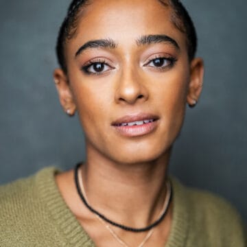 A person with braided hair, natural makeup, and wearing a green sweater and layered necklaces, looking directly at the camera.