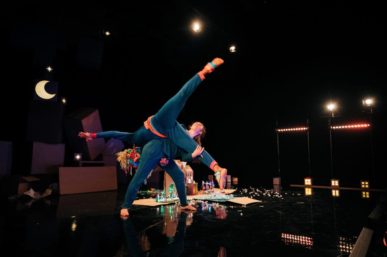 Two performers in blue costumes execute an acrobatic lift on stage, with set decorations including a crescent moon and scattered props in the background.
