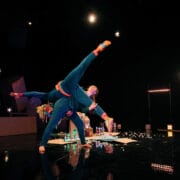 Two performers in blue costumes execute an acrobatic lift on stage, with set decorations including a crescent moon and scattered props in the background.
