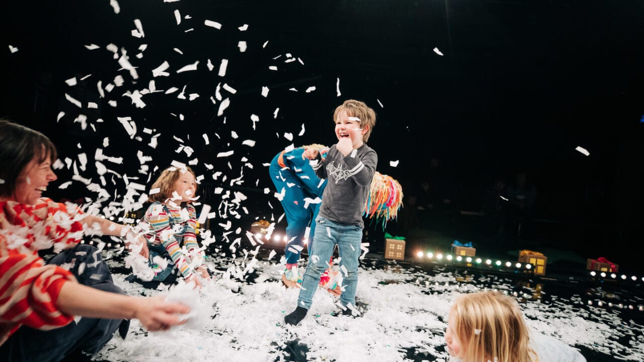 Children play and laugh as white paper confetti falls around them on a stage, with adults joining in the activity.