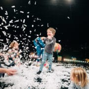 Children play and laugh as white paper confetti falls around them on a stage, with adults joining in the activity.