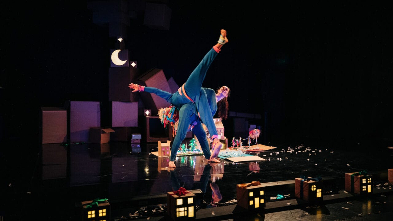 Two performers in colorful costumes do an acrobatic pose on stage, surrounded by cardboard props and small illuminated house models, under dim lighting with a crescent moon backdrop.