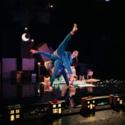 Two performers in colorful costumes do an acrobatic pose on stage, surrounded by cardboard props and small illuminated house models, under dim lighting with a crescent moon backdrop.