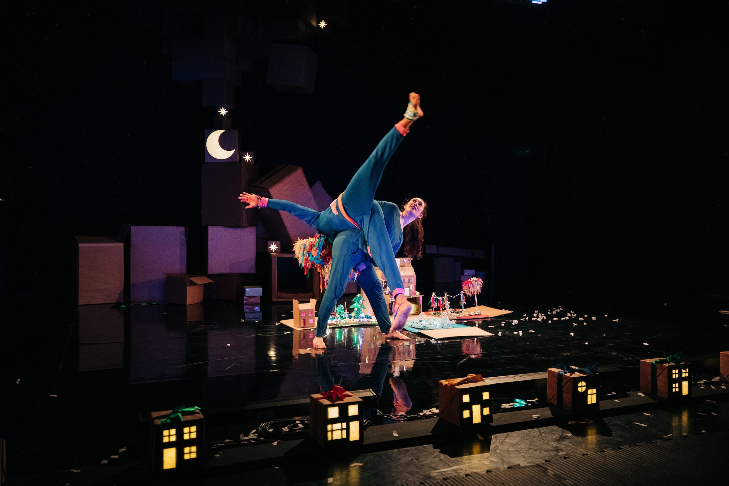 Two performers in colorful costumes do an acrobatic pose on stage, surrounded by cardboard props and small illuminated house models, under dim lighting with a crescent moon backdrop.