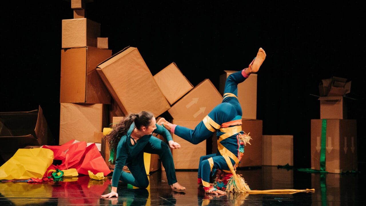 Two performers in colorful costumes pose onstage; one balances on their head while the other kneels beside them. Large cardboard boxes and colorful paper are in the background.