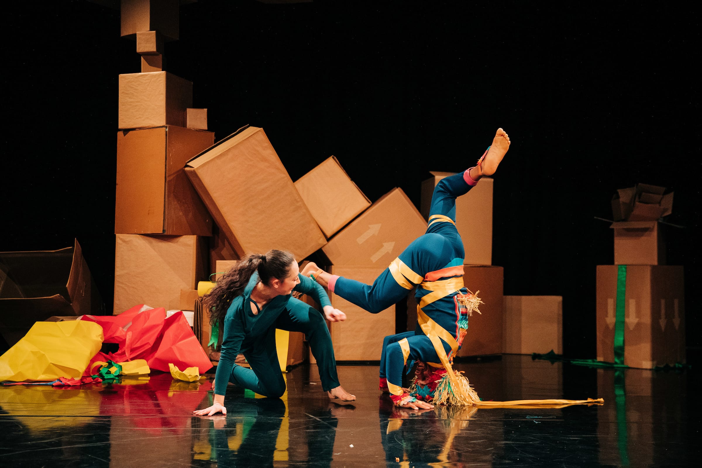 Two performers in colorful costumes pose onstage; one balances on their head while the other kneels beside them. Large cardboard boxes and colorful paper are in the background.