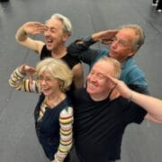 Four adults stand in a staggered formation indoors at Festival Theatre, all smiling and giving a salute gesture while looking upwards, embodying the spirit of the High Life.
