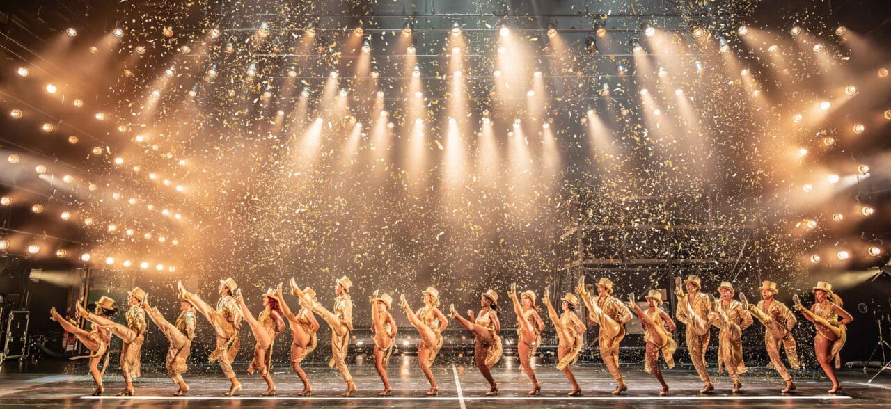 A line of performers in gold costumes and hats do a synchronized high kick on stage under bright lights with gold confetti falling.
