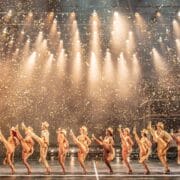 A line of performers in gold costumes and hats do a synchronized high kick on stage under bright lights with gold confetti falling.