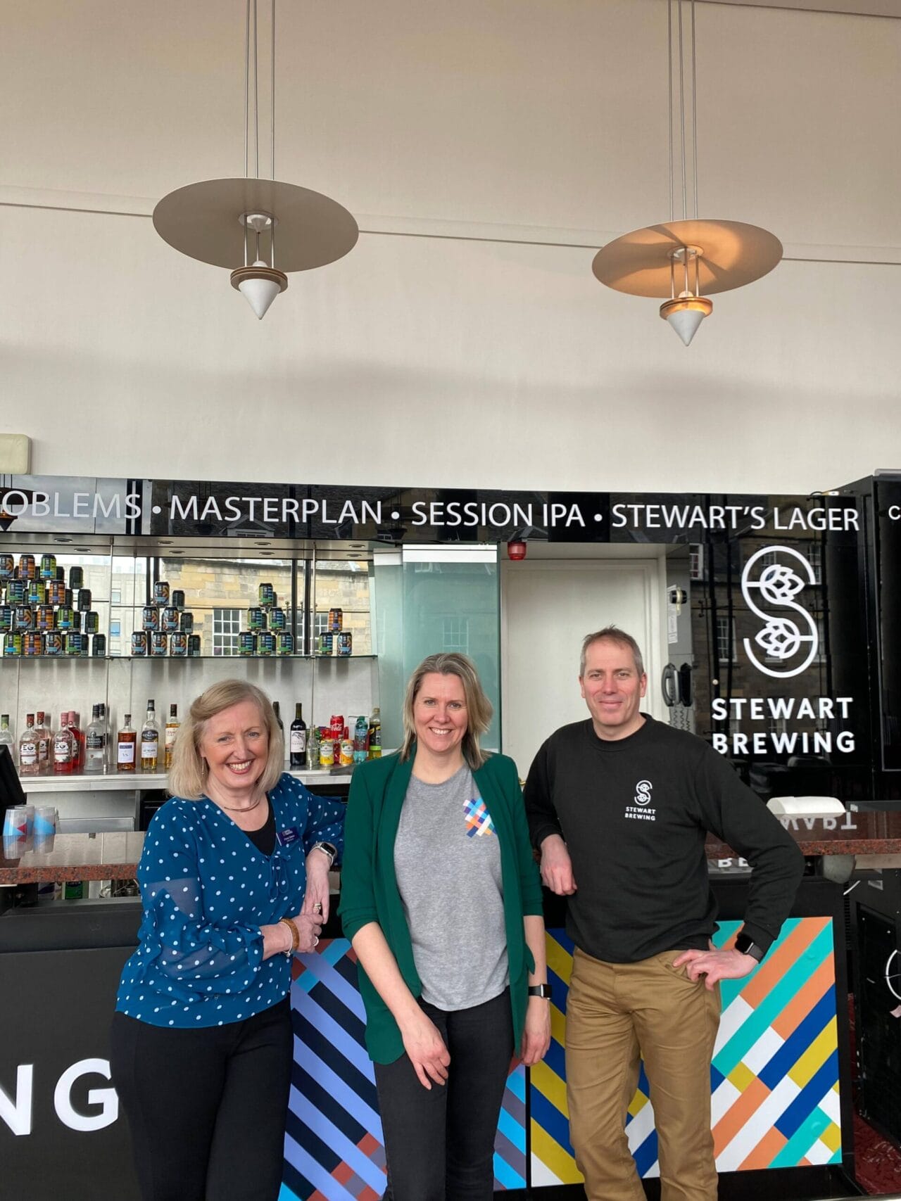 Three people stand and smile in front of a bar with Stewart Brewing signage and a colorful counter design in a well-lit indoor space.