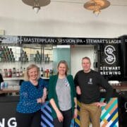 Three people stand and smile in front of a bar with Stewart Brewing signage and a colorful counter design in a well-lit indoor space.