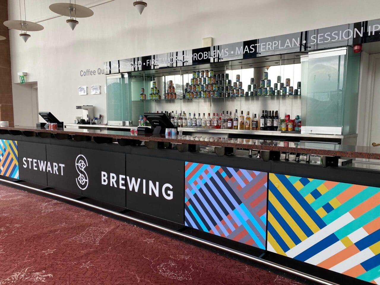 A modern bar counter with Stewart Brewing signage, colorful geometric panels, and shelves of bottles and cans behind the counter.