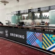 A modern bar counter with Stewart Brewing signage, colorful geometric panels, and shelves of bottles and cans behind the counter.