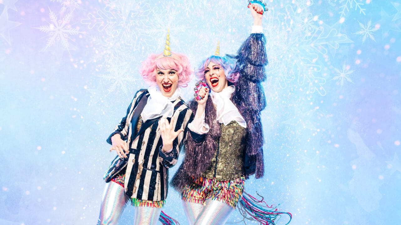 Two people dressed in colorful unicorn-themed costumes with pink and purple wigs, striped jackets, and sparkly shorts stand smiling and posing against a blue, snowy, festive background.