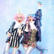 Two people dressed in colorful unicorn-themed costumes with pink and purple wigs, striped jackets, and sparkly shorts stand smiling and posing against a blue, snowy, festive background.