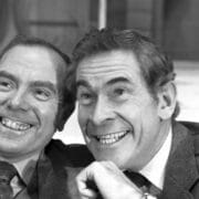 Two men in suits smiling and posing closely together for the camera in a black and white photograph.