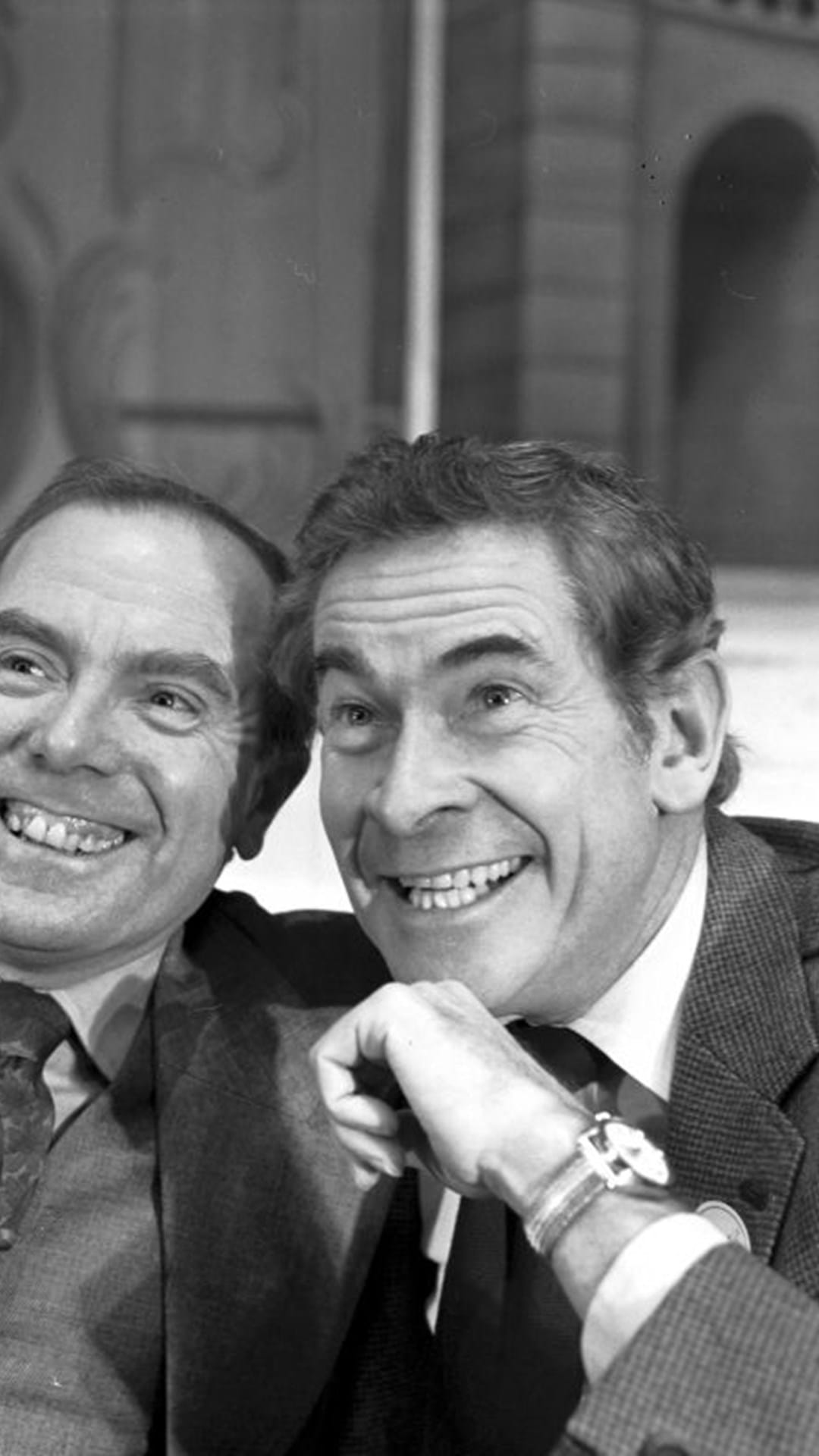 Two men in suits smiling and posing closely together for the camera in a black and white photograph.