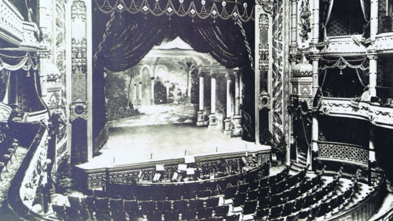 Black and white photo of an ornate, empty theater with a stage set resembling a classical garden, rows of seats, and a decorative proscenium arch.