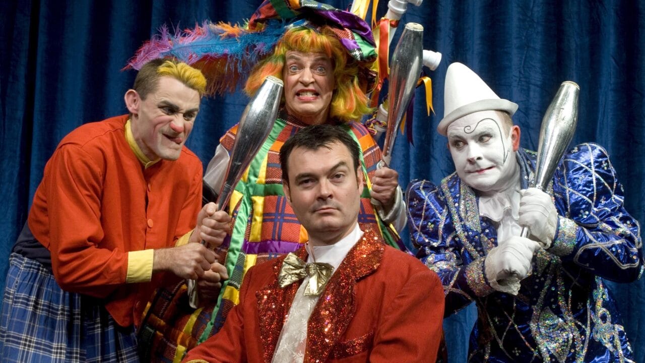 Four people dressed as clowns in colorful costumes pose with juggling clubs against a blue curtain backdrop.