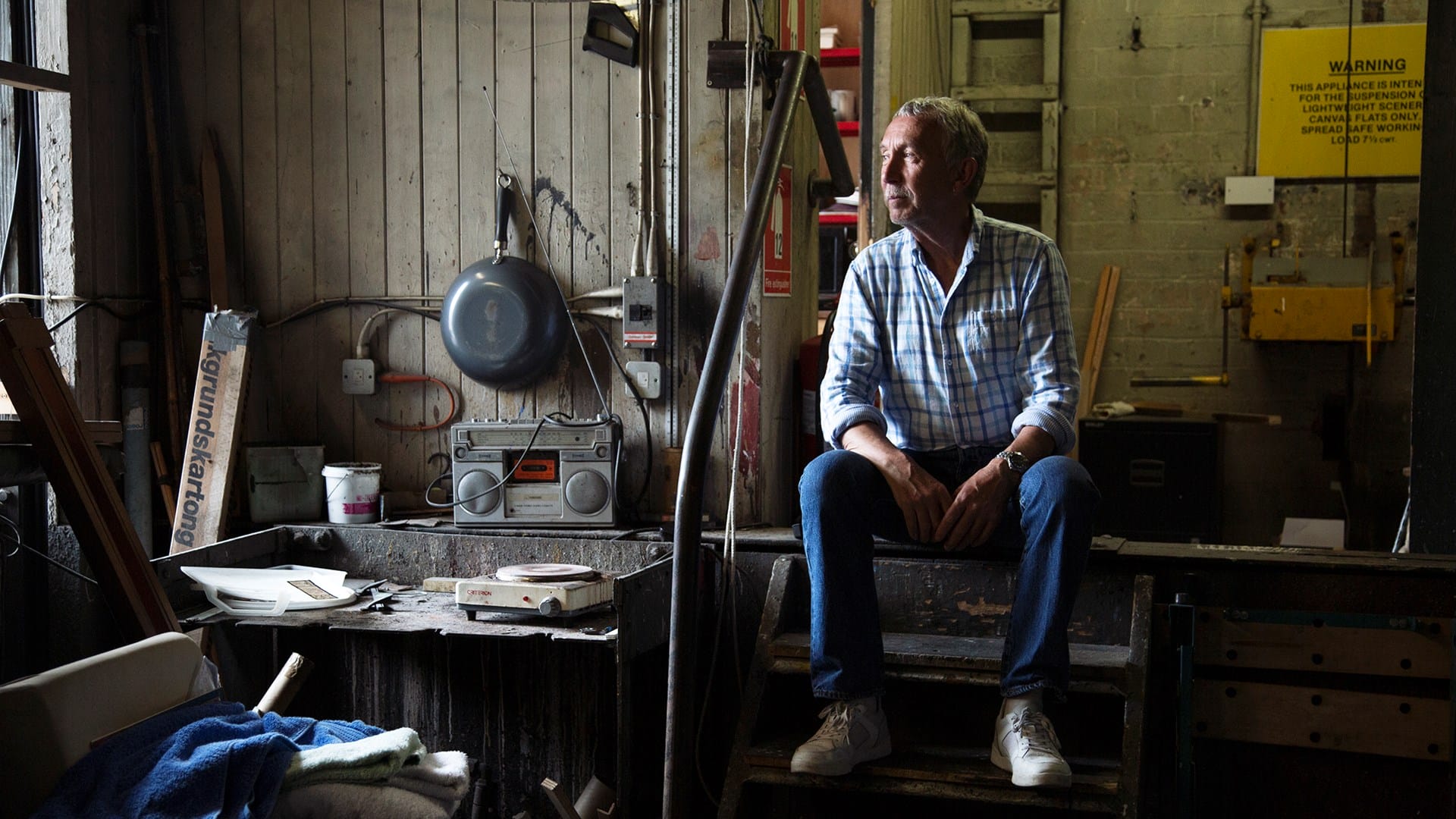 A man in a plaid shirt and jeans sits on steps inside a cluttered workshop, with tools, equipment, and a radio visible around him.
