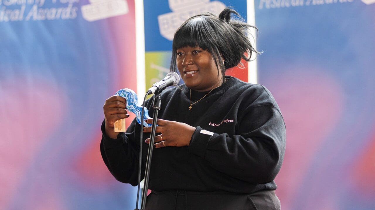 A person speaks into a microphone while holding a blue object and an envelope, standing in front of a colorful backdrop with text.