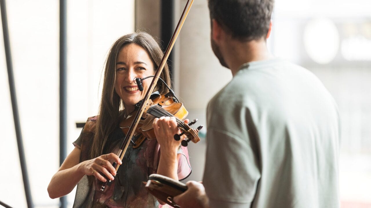 A woman plays the violin and smiles at a man holding a small notebook or device, both indoors in a well-lit area.