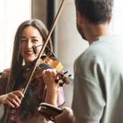 A woman plays the violin and smiles at a man holding a small notebook or device, both indoors in a well-lit area.