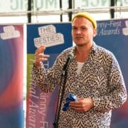 A man in a patterned shirt and yellow beanie speaks into a microphone at The Skinny-Fest Festival Awards, standing in front of event banners.