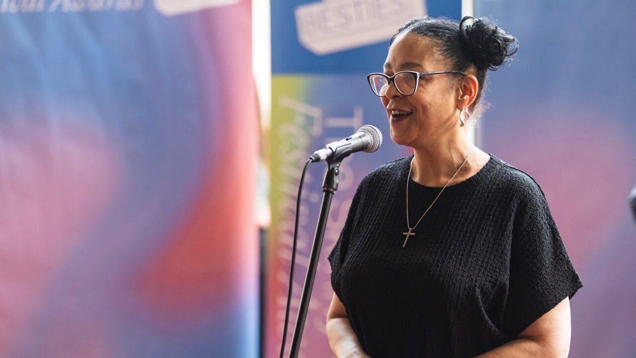 A woman wearing glasses and a black top speaks into a microphone, standing in front of colorful, blurred banners.