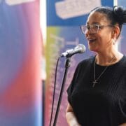 A woman wearing glasses and a black top speaks into a microphone, standing in front of colorful, blurred banners.