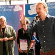 A man speaks into a microphone at an awards event while others stand behind him, with "The Skinny-Fest Festival Awards" banners in the background.