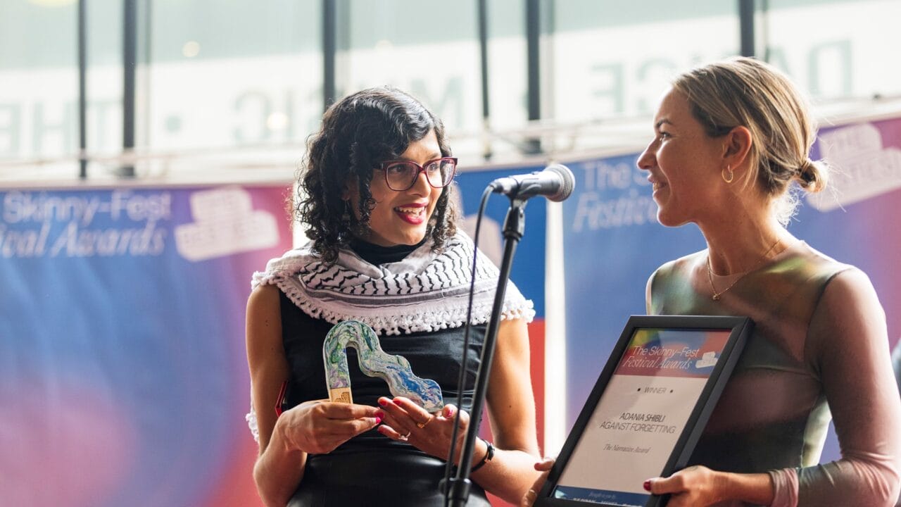 Two women stand at a microphone; one holds a glass award and smiles, while the other presents a framed certificate at an awards event.