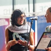 Two women stand at a microphone; one holds a glass award and smiles, while the other presents a framed certificate at an awards event.