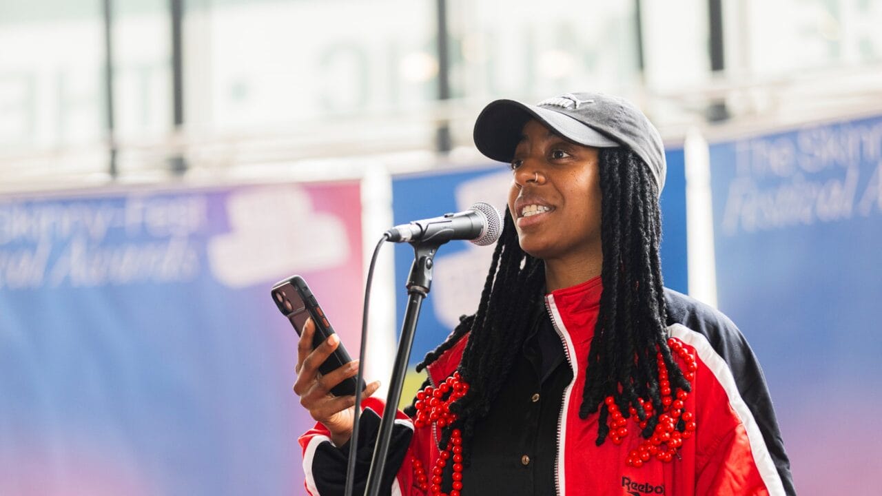 A person in a red and black jacket speaks into a microphone while holding a smartphone, standing in front of a blurred banner.