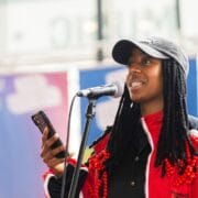 A person in a red and black jacket speaks into a microphone while holding a smartphone, standing in front of a blurred banner.