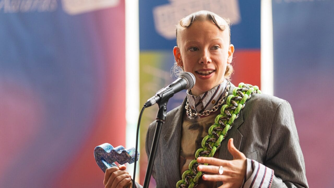 A person wearing a striped suit and chunky necklace speaks into a microphone, holding a blue heart-shaped award.