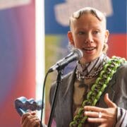 A person wearing a striped suit and chunky necklace speaks into a microphone, holding a blue heart-shaped award.