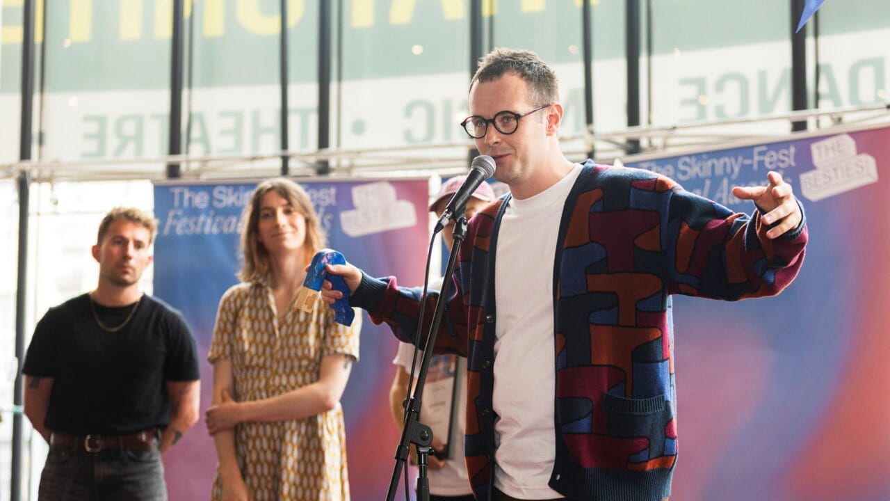 A man in glasses speaks into a microphone on stage, holding an object, with two people standing in the background.