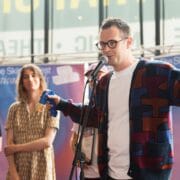 A man in glasses speaks into a microphone on stage, holding an object, with two people standing in the background.