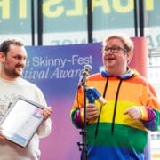Two people stand indoors at an awards event; one holds a framed certificate, the other holds a trophy and speaks into a microphone, wearing a rainbow-striped hoodie.