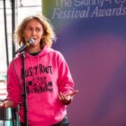 A person in a pink "Pussy Riot" hoodie speaks into a microphone on stage at The Skinny Fest Festival Awards event.