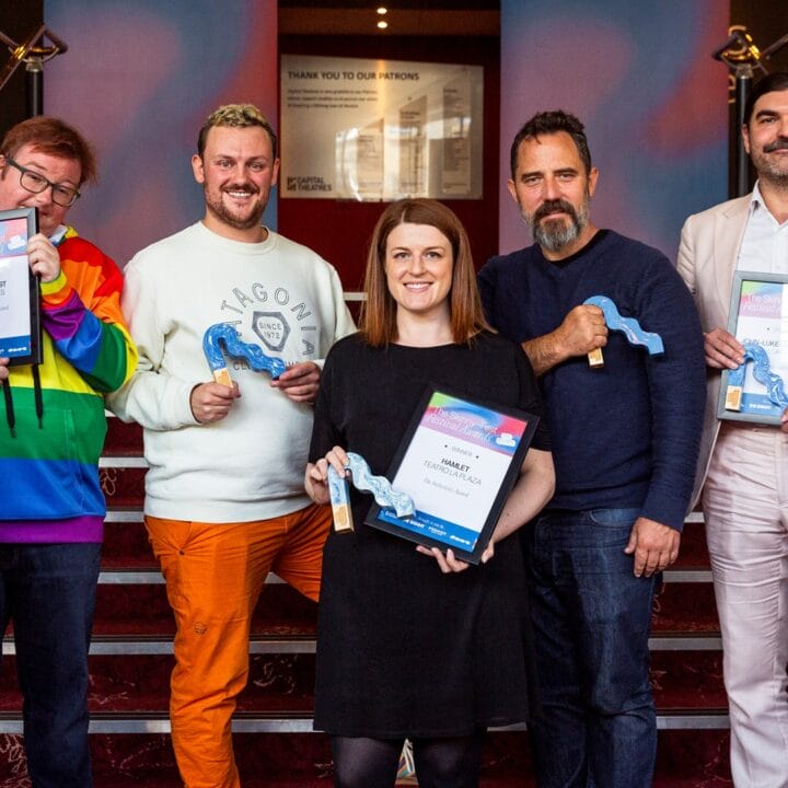 Seven people stand in a row indoors, holding certificates and blue ribbon-shaped awards, posing and smiling for the camera on a staircase.