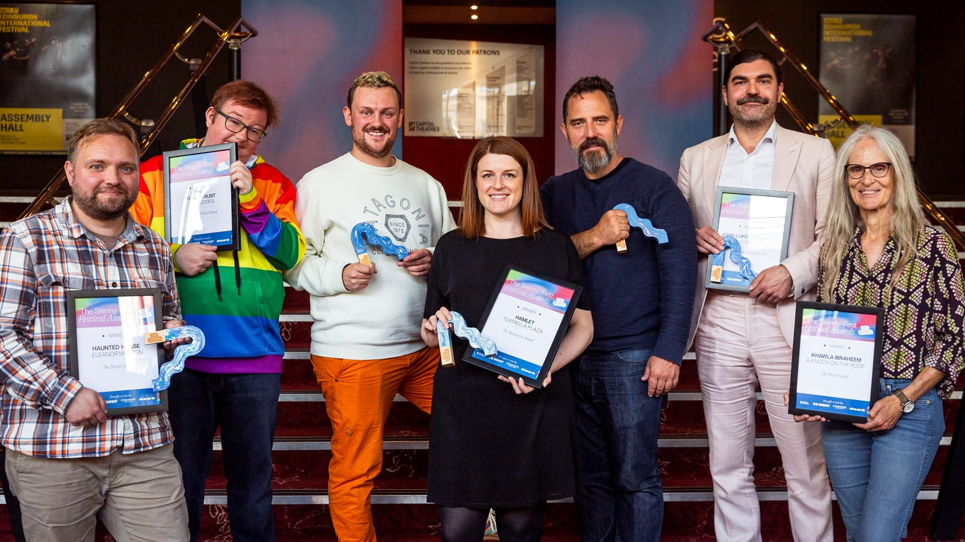 Seven people stand in a row indoors, holding certificates and blue ribbon-shaped awards, posing and smiling for the camera on a staircase.