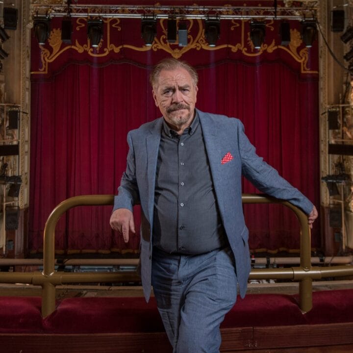 A man in a blue suit stands in a theatre balcony with red velvet seats, ornate lighting, and a large red curtain on stage in the background.