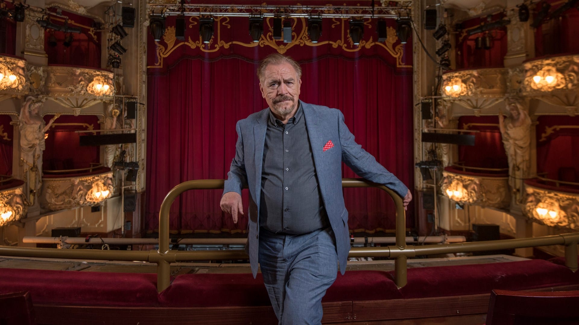 A man in a blue suit stands in a theatre balcony with red velvet seats, ornate lighting, and a large red curtain on stage in the background.